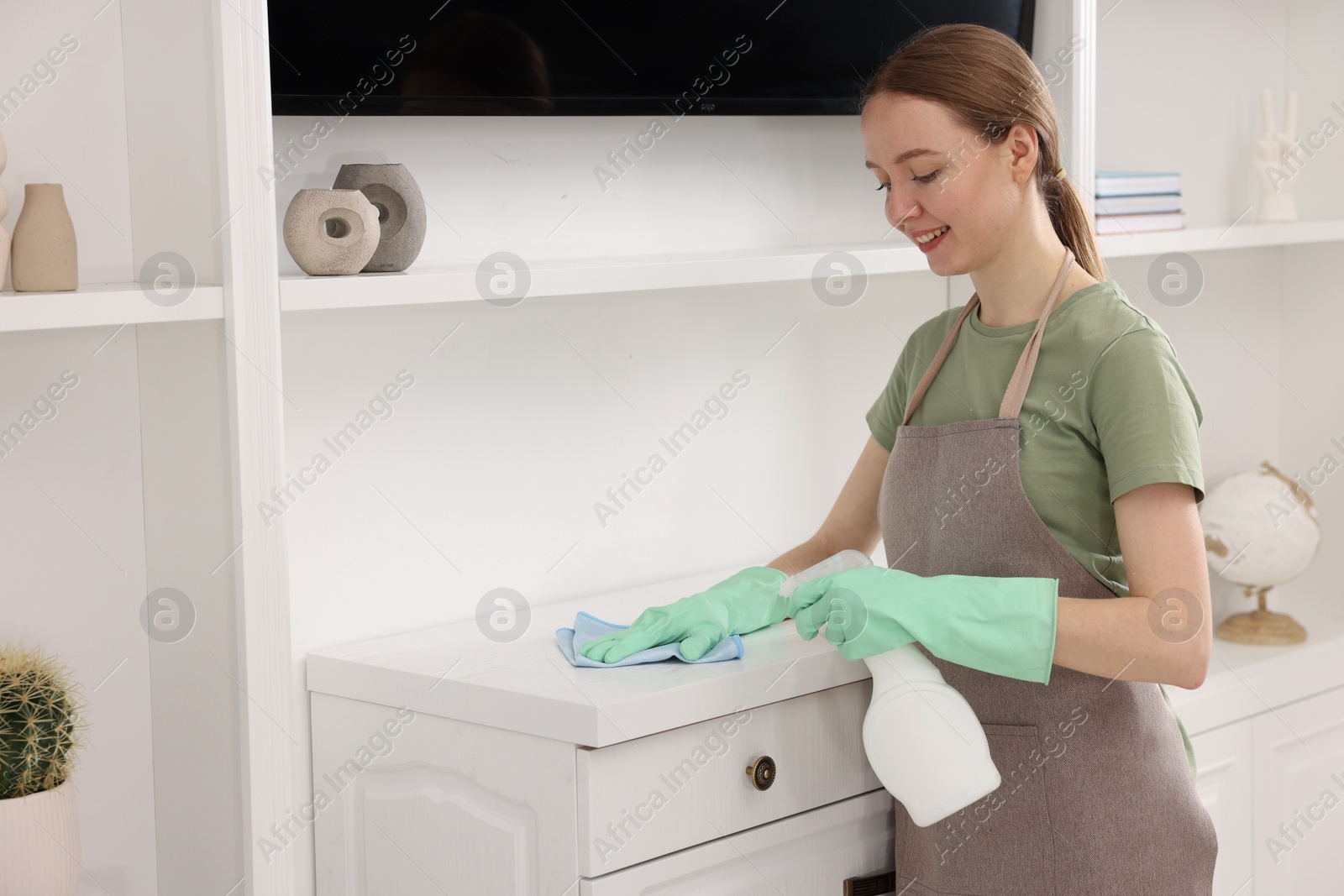 Photo of Woman with spray bottle and microfiber cloth cleaning white chest of drawers in room,