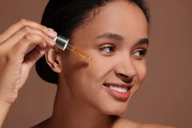 Smiling woman applying serum onto her face on brown background, closeup