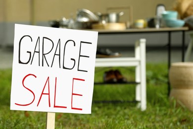 Photo of Sign Garage sale written on cardboard near tables with different stuff in yard, closeup. Space for text