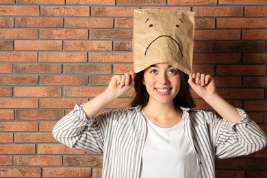 Photo of Happy woman wearing paper bag with drawn sad face near brick wall