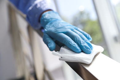 Photo of Woman in latex gloves cleaning railing with wet wipe indoors, closeup