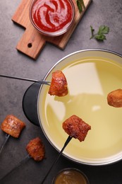 Oil in fondue pot, forks with fried meat pieces and sauces on grey table, flat lay