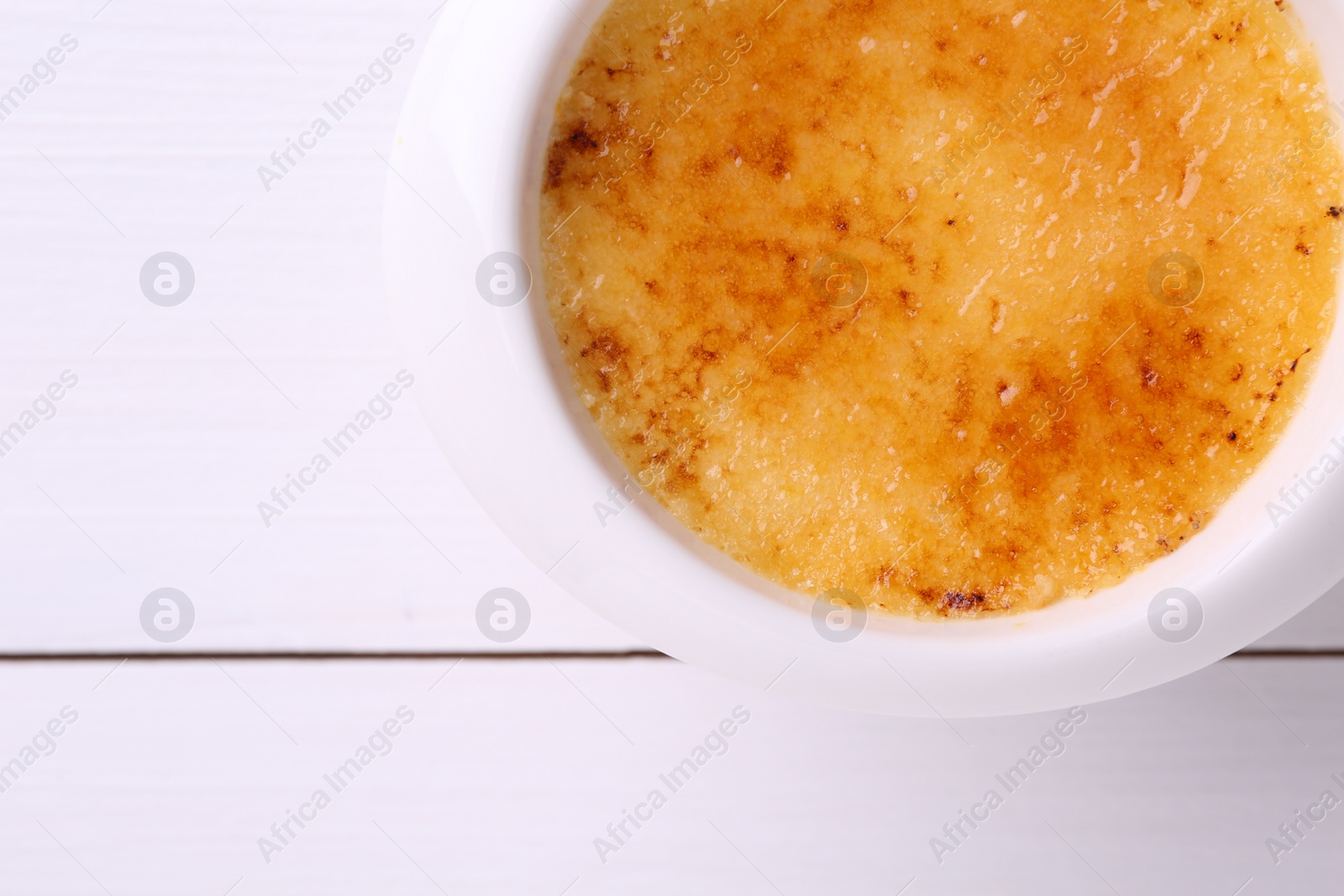 Photo of Delicious creme brulee in bowl on white wooden table, top view. Space for text