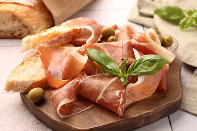 Slices of tasty cured ham, olives, bread and basil on tiled table, closeup