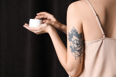 Photo of Woman with tattoo holding jar of cream against dark background, closeup