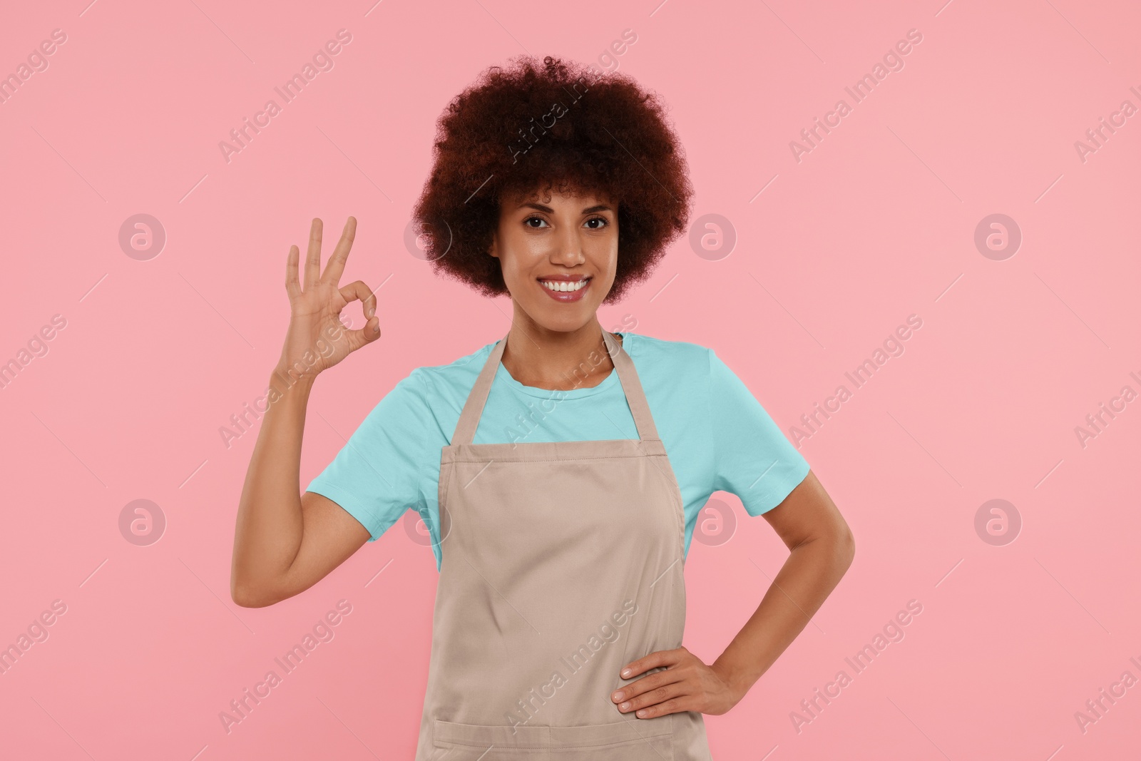 Photo of Happy young woman in apron showing ok gesture on pink background
