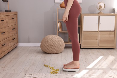 Woman standing on floor scale and measuring tape at home, closeup. Weight control