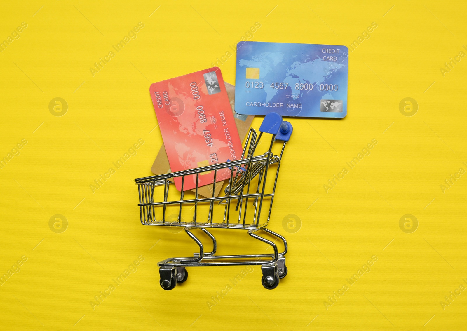 Photo of Small metal shopping cart and credit cards on yellow background, top view