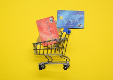 Small metal shopping cart and credit cards on yellow background, top view