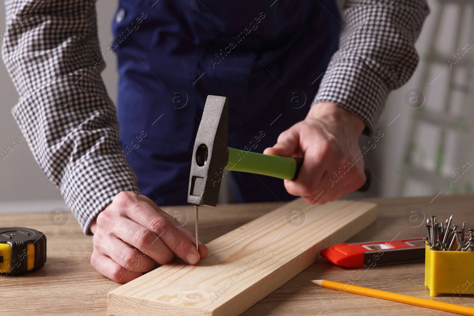 Photo of Professional repairman hammering nail into board at wooden table indoors, closeup