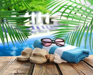 Beach towel, sunglasses, shoes and sea shell on wooden surface near outdoor swimming pool