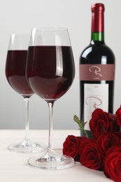 Bottle and glasses of red wine near beautiful roses on white wooden table