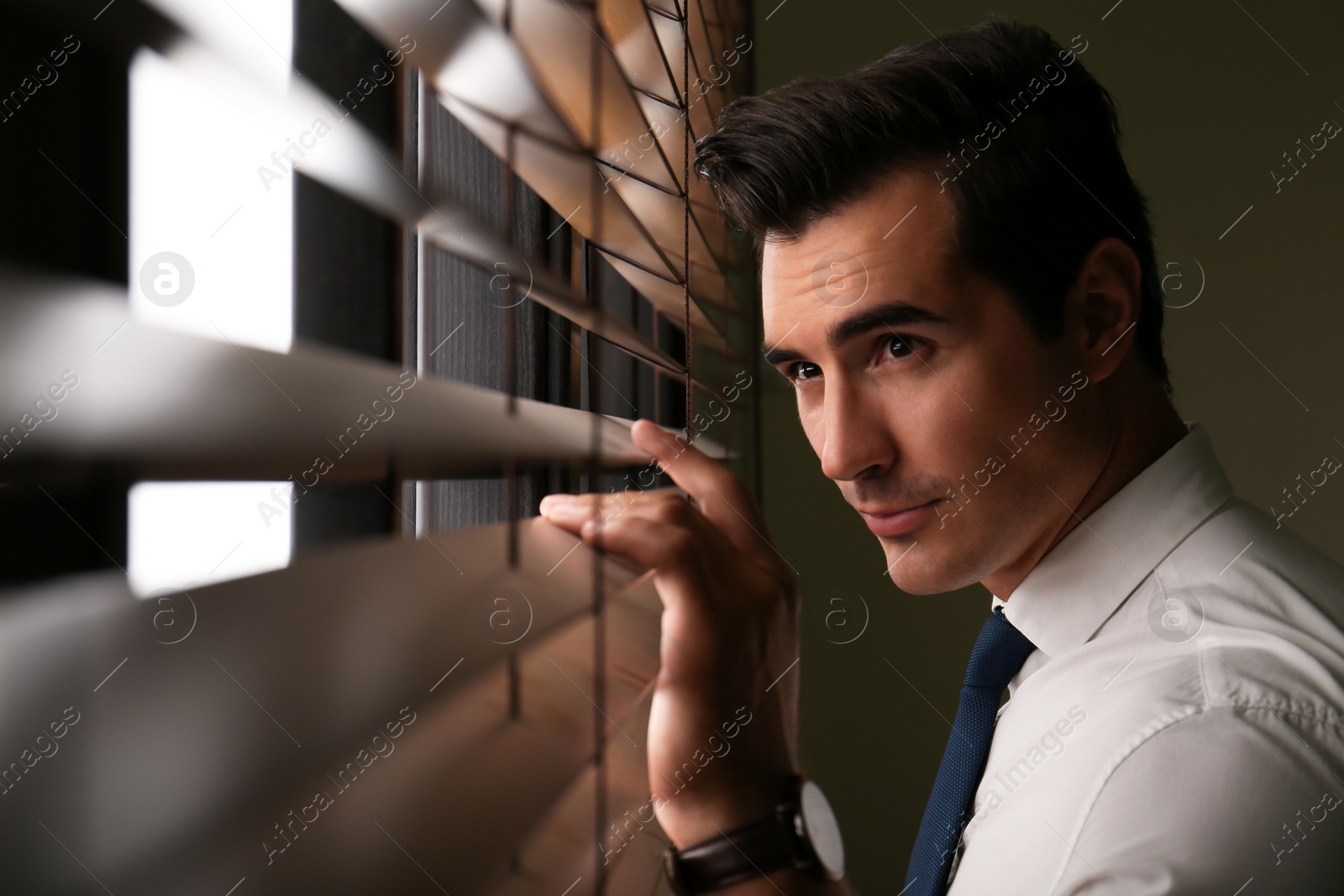 Photo of Handsome young man looking through window blinds indoors