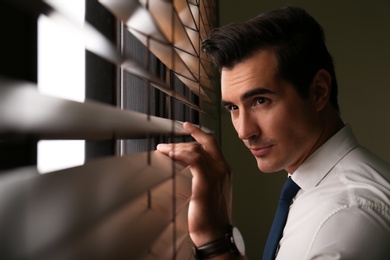 Photo of Handsome young man looking through window blinds indoors