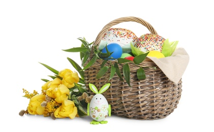 Photo of Basket with delicious Easter cakes, dyed eggs and flowers on white background