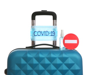 Photo of Antiseptic spray, stop sign and protective mask with inscription COVID-19 on suitcase against white background. Travel restriction during coronavirus pandemic