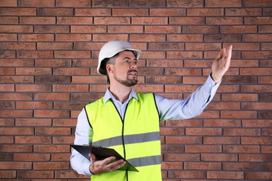 Photo of Male industrial engineer in uniform with clipboard on brick wall background. Safety equipment