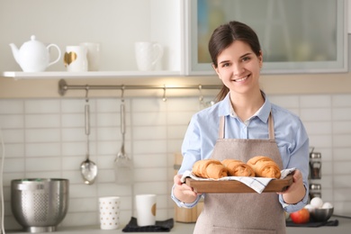 Woman holding wooden tray with delicious croissants in kitchen