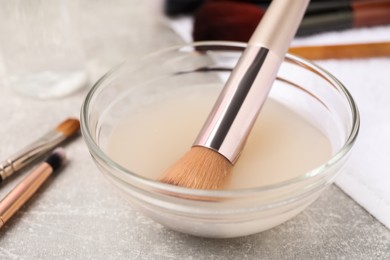 Photo of Makeup brush in bowl with cleanser on grey table, closeup