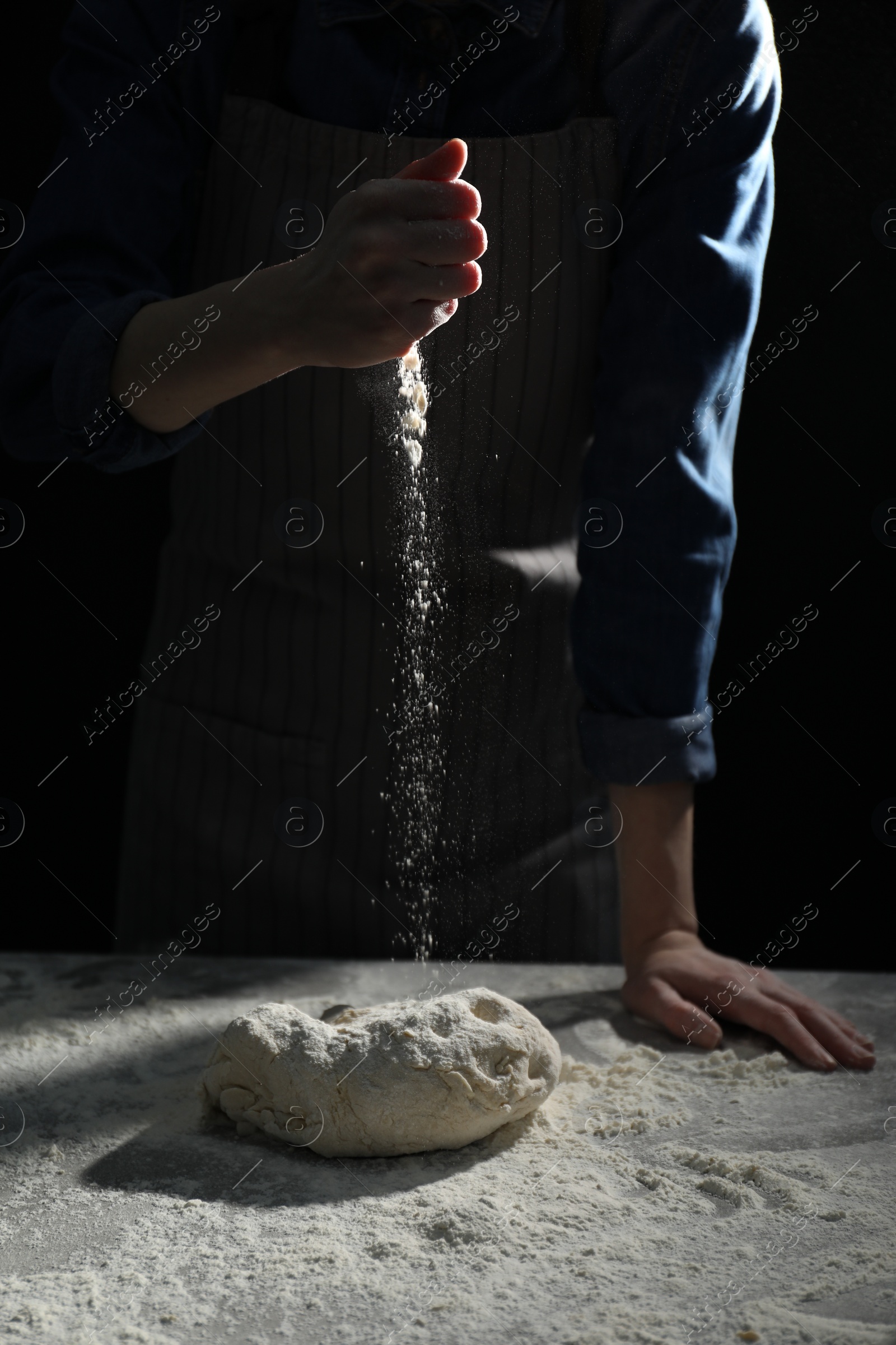 Photo of Making bread. Woman sprinkling flour over dough at table on dark background, closeup