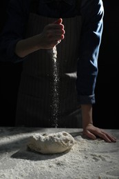 Photo of Making bread. Woman sprinkling flour over dough at table on dark background, closeup