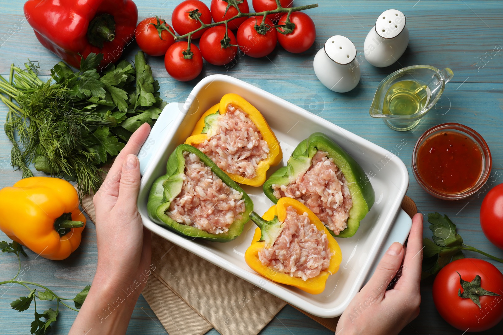 Photo of Woman holding dish with raw stuffed peppers at light blue wooden table, top view