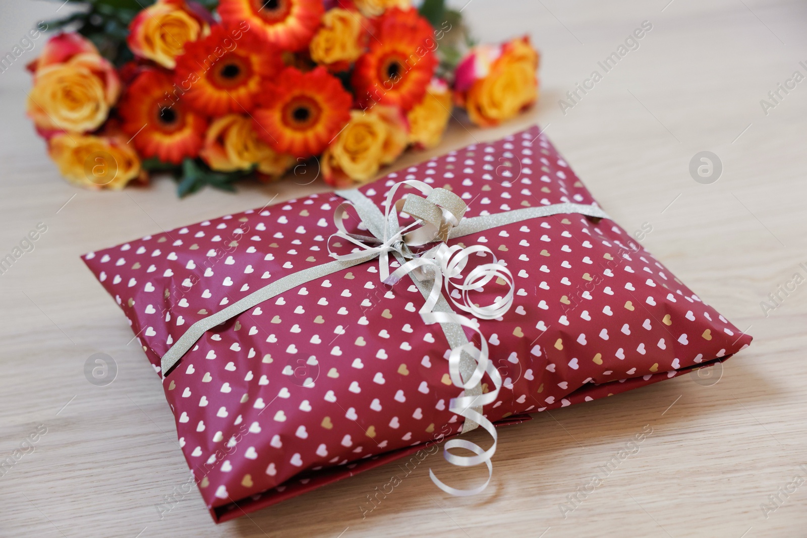 Photo of Parcel wrapped in heart patterned paper and beautiful flowers on wooden table, closeup