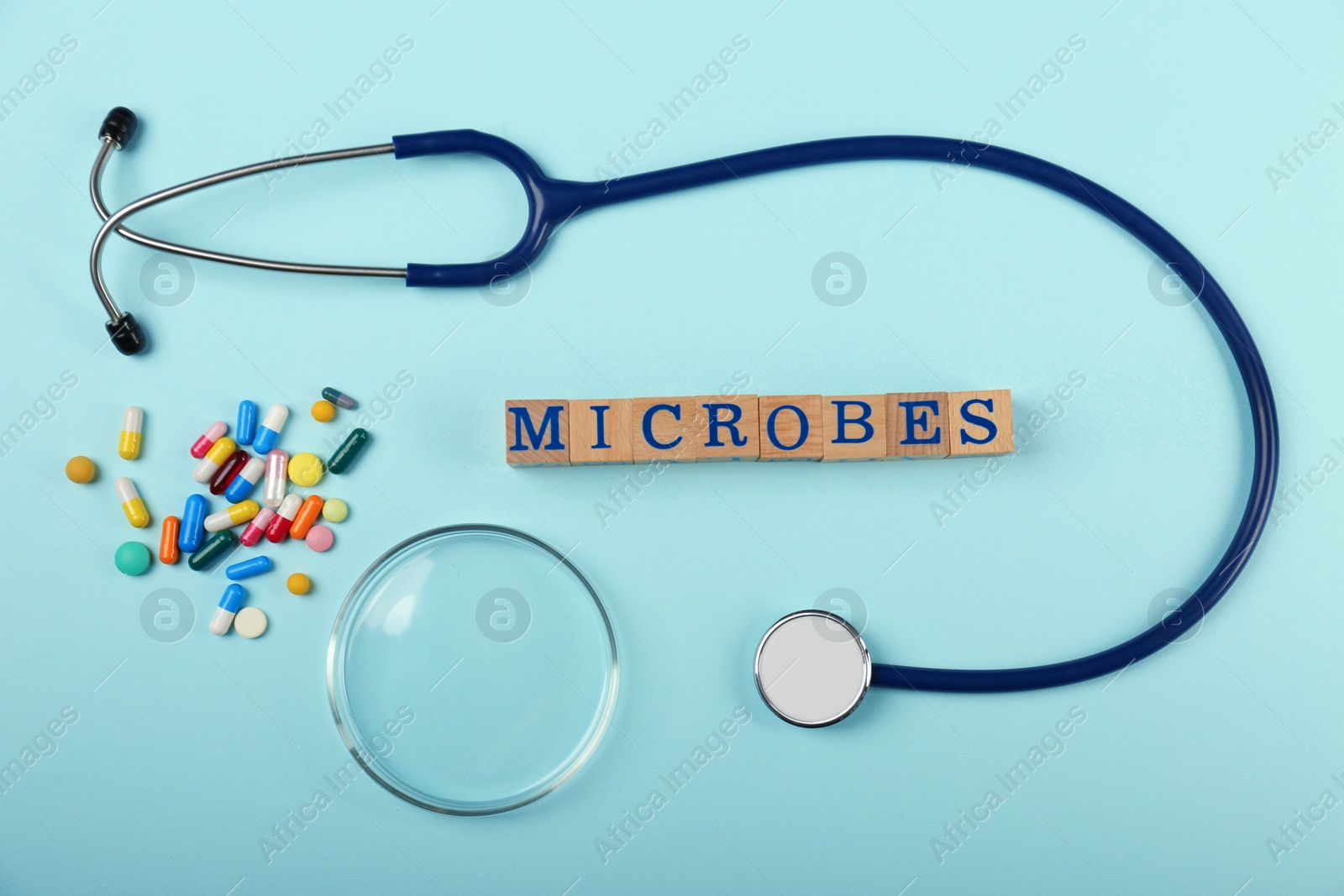 Photo of Word Microbes made with wooden cubes, pills and stethoscope on light blue background, flat lay