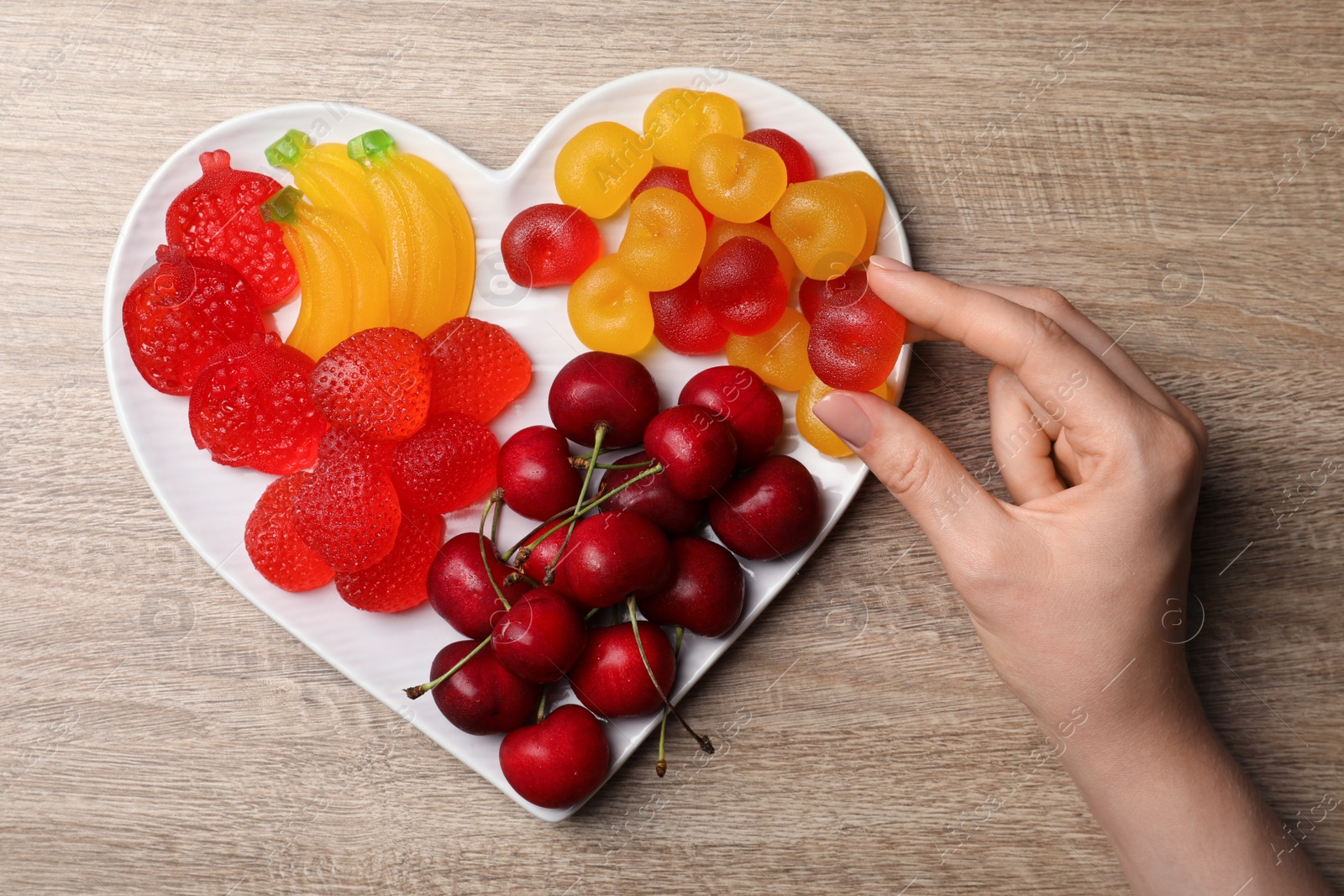 Photo of Woman taking gummy cherry candy from plate on wooden table, top view