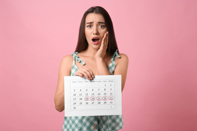 Young woman holding calendar with marked menstrual cycle days on pink background