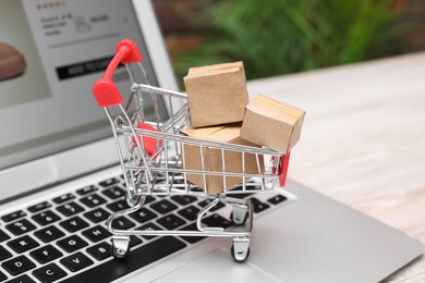 Photo of Internet store. Small cardboard boxes, shopping cart and laptop on light wooden table, closeup