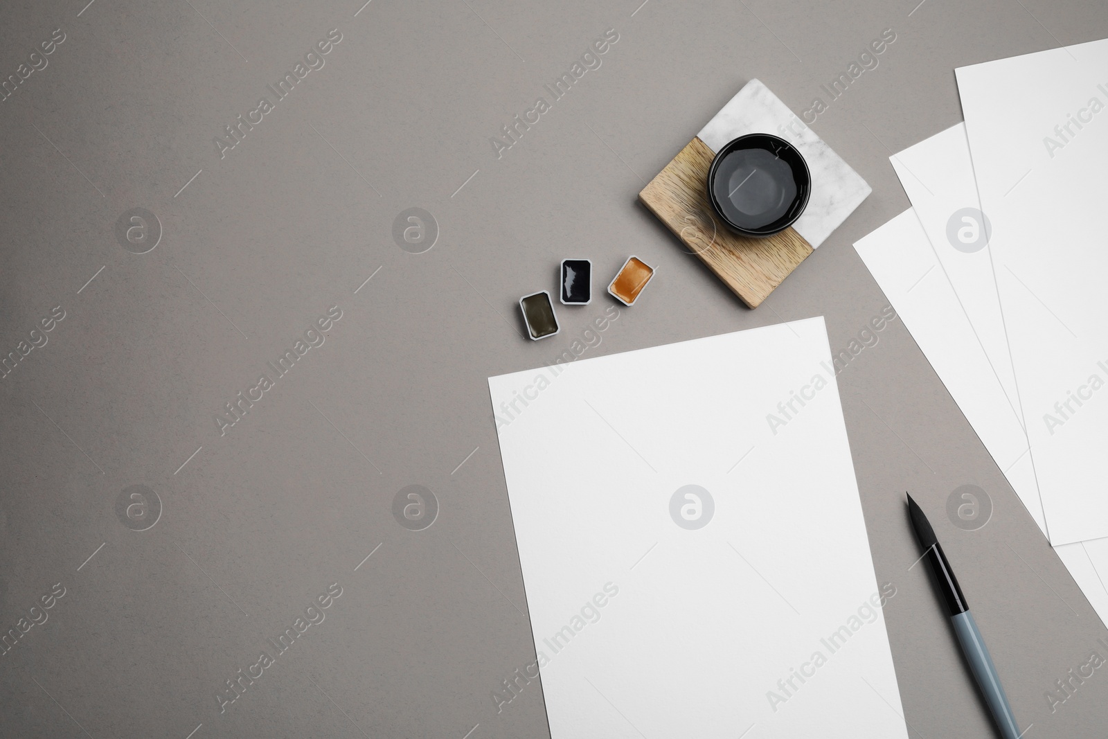 Photo of Blank sheets of paper, watercolor paints and brush on grey table, flat lay. Space for text
