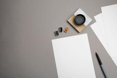 Blank sheets of paper, watercolor paints and brush on grey table, flat lay. Space for text