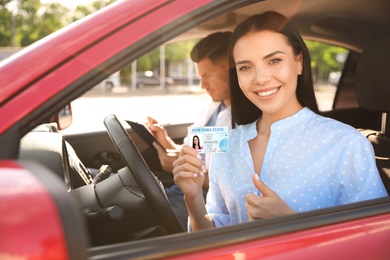 Photo of Young woman holding license while sitting in car with instructor. Driving school