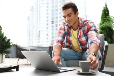 Portrait of handsome young African-American man with laptop and cup of drink in outdoor cafe