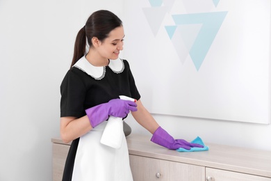 Young maid cleaning furniture with rag in hotel room