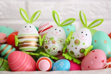 Photo of Three eggs as Easter bunnies among painted others near white brick wall, closeup