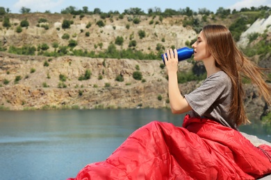 Image of Young camper drinking from bottle in sleeping bag near lake