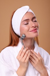 Photo of Young woman massaging her face with metal roller on pale orange background