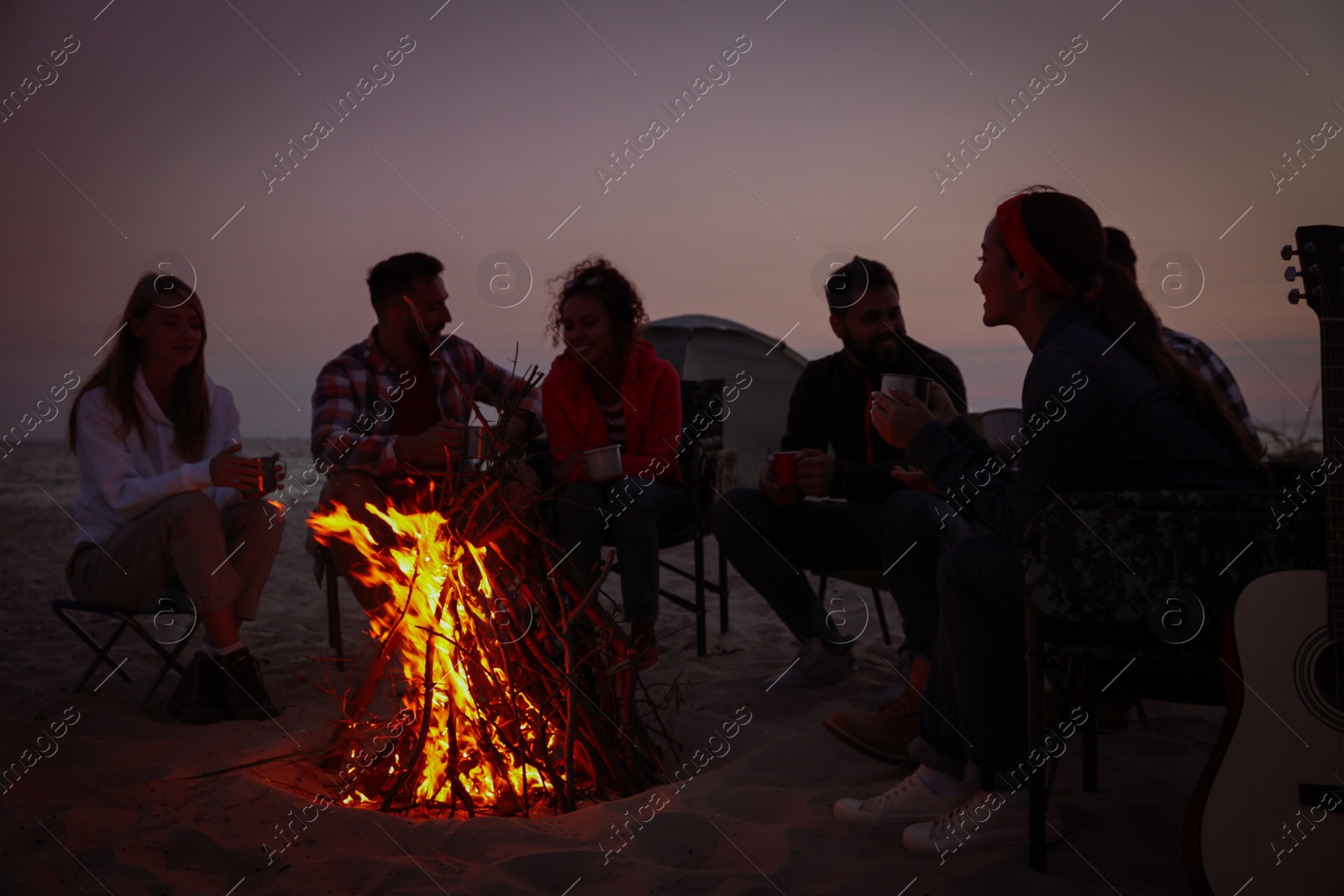 Photo of Group of friends gathering around bonfire on beach in evening. Camping season