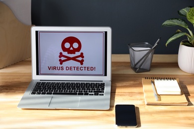 Photo of Smartphone, stationery and laptop with warning about virus attack on wooden table indoors