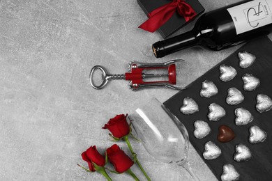 Bottle of red wine, glass, heart shaped chocolate candies, corkscrew, roses and gift box on light grey textured table, flat lay. Space for text