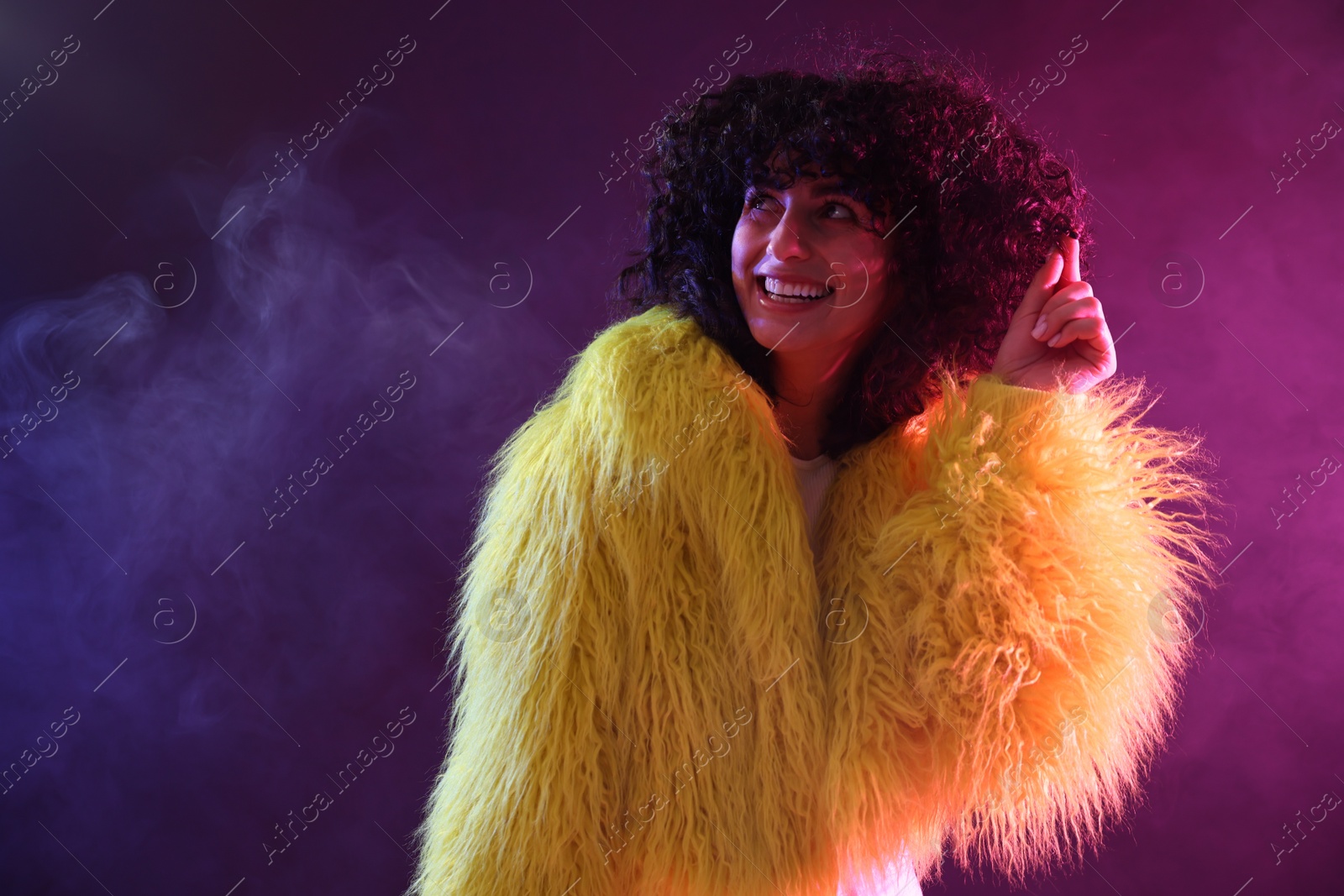 Photo of Beautiful young woman in yellow fur coat on color background in neon lights and smoke