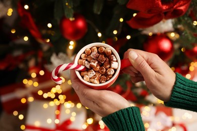Photo of Woman holding cup of delicious cocoa with marshmallows and candy cane near decorated Christmas tree, above view