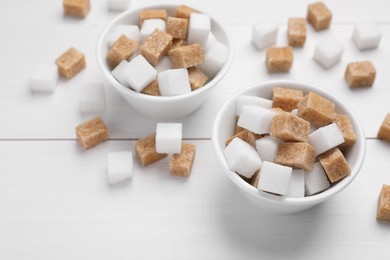 Different sugar cubes in bowls on white wooden table, above view