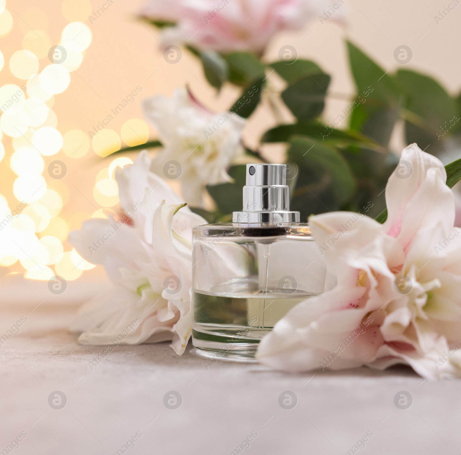 Photo of Bottle of perfume and beautiful lily flowers on table against beige background with blurred lights, closeup