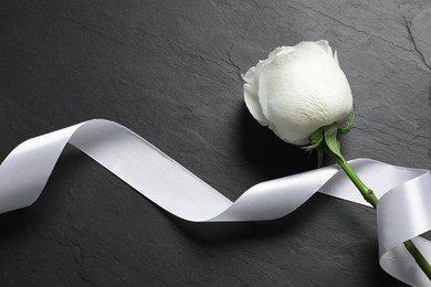 Photo of White rose and ribbon on black table, top view. Funeral symbols