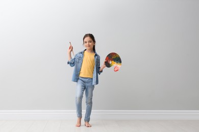 Photo of Little girl with brush and paint palette near light grey wall indoors