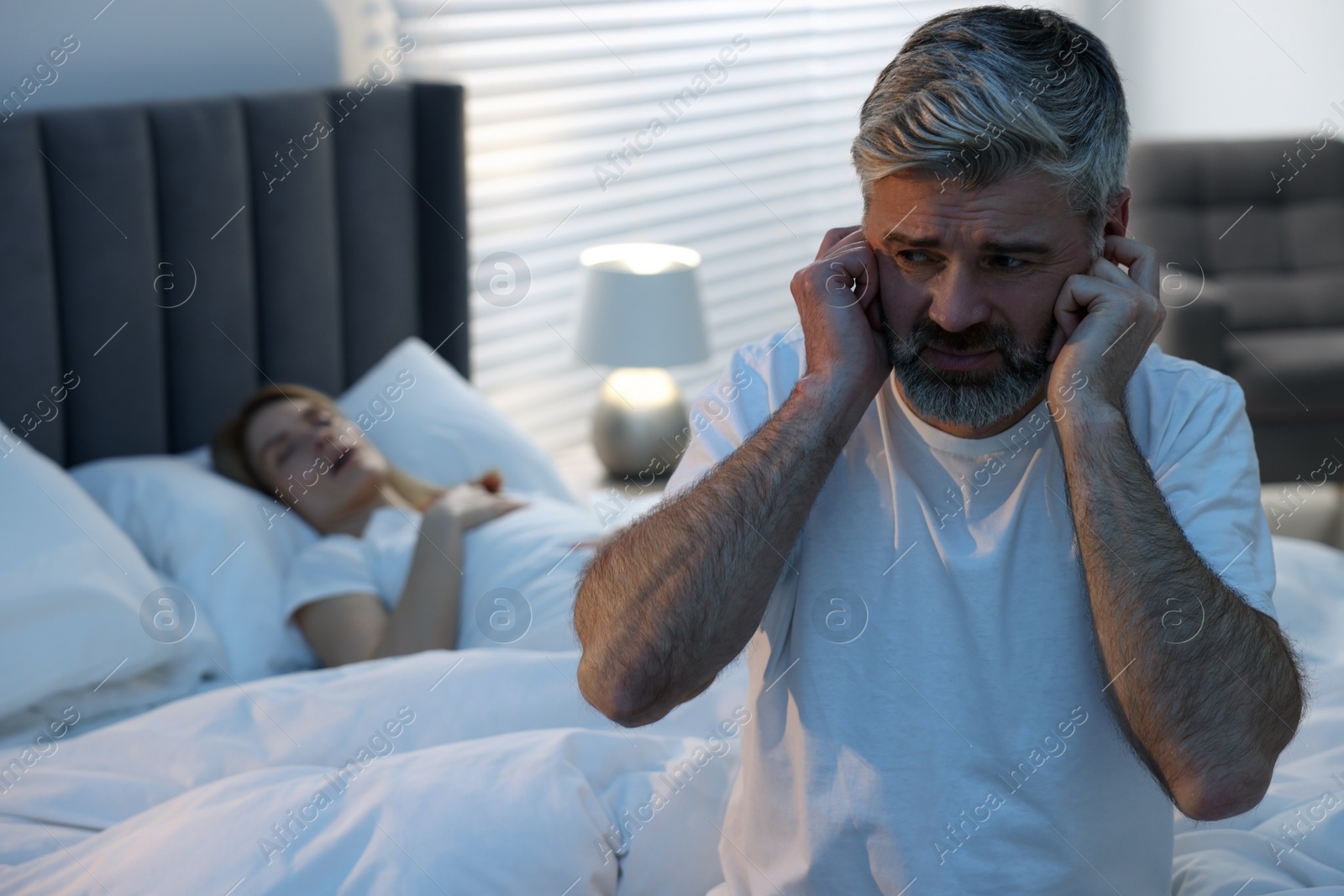 Photo of Irritated man covering his ears in bed at home, space for text. Problem with snoring wife