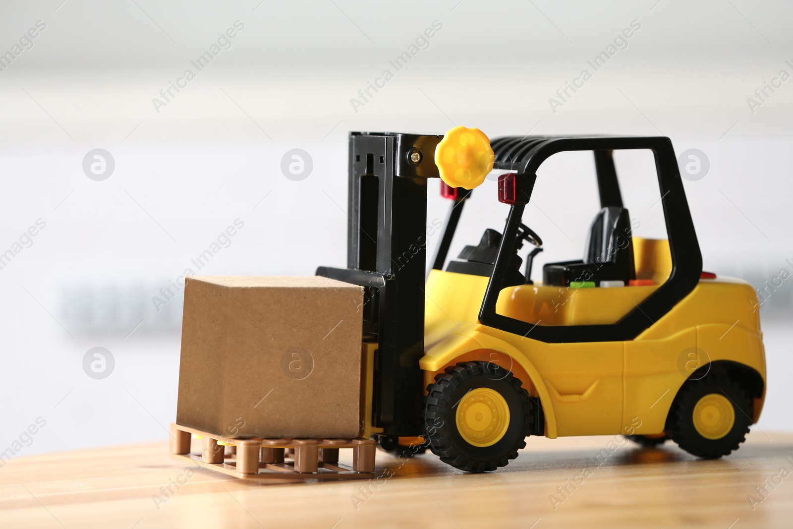 Photo of Toy forklift with cardboard box on table against blurred background. Courier service